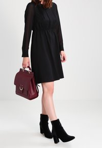 Svart klänning med långa transparenta ärmar och plisséer vid halsen, kombinerad med svarta klackade ankelstövlar och en mörkröd läderhandväska.