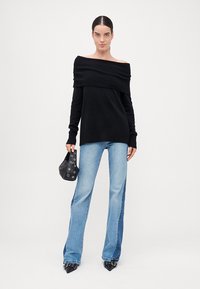 Schwarzer, schulterfreier, gerippter Pullover mit langen Ärmeln, kombiniert mit hellblauen Schlagjeans. Die Accessoires umfassen eine kleine schwarze Tasche und spitze Schuhe.