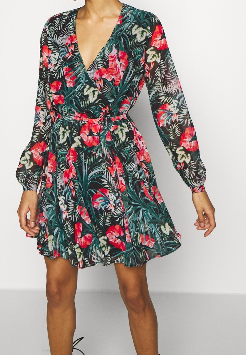 Femme portant une robe à manches longues, col en V, longueur jusqu'aux genoux, avec un motif coloré de fleurs et de feuilles vertes sur fond noir.