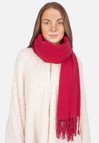 Roter Wollschal mit fransigen Kanten, um den Hals drapiert, kombiniert mit einem strukturierten cremefarbenen Cardigan. Glatter Stoff, warme Ausstrahlung.