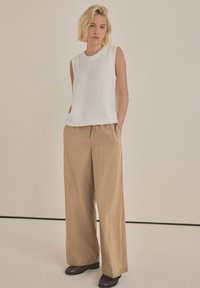 Maglione bianco senza maniche con scollatura a costine, abbinato a pantaloni a gamba larga beige a righe sottili. Il modello indossa scarpe marroni scure, con le mani nelle tasche.