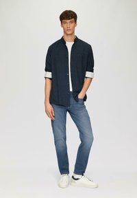 Chemise bleu foncé à manches retroussées, t-shirt blanc en dessous, jean bleu clair et baskets blanches avec accents marine.