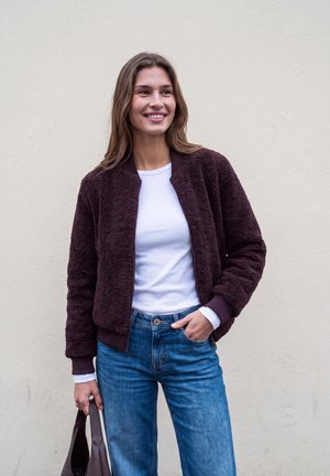 Jeune femme souriante, portant une veste marron foncé, une chemise blanche, un jean, tenant un sac foncé devant un mur clair uni.