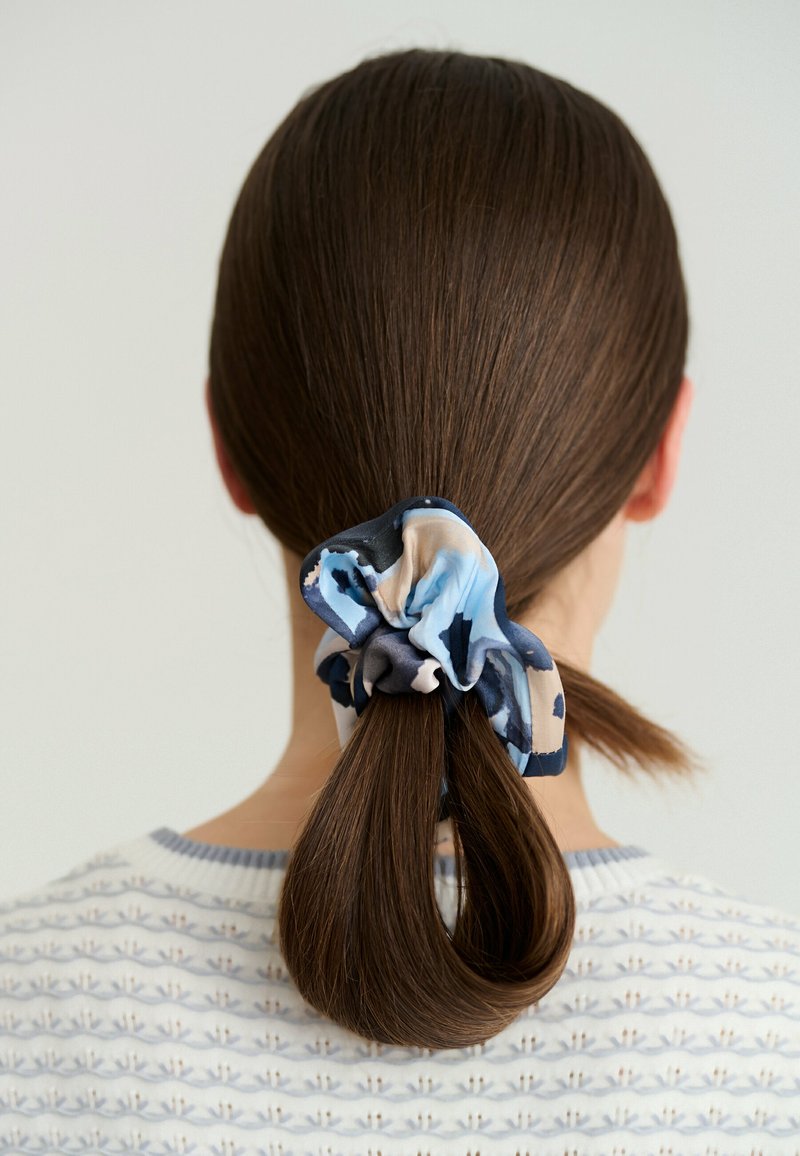 Cheveux bruns attachés en une queue de cheval basse avec un chouchou à motifs bleu, noir et beige, portant un pull blanc à motifs.