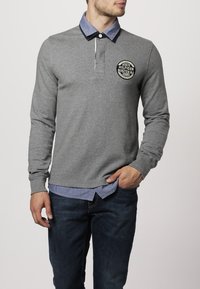 Graues Langarm-Polohemd mit einem Denim-Kragen, das ein rundes Logo auf der Brust und eine taillierte Passform aufweist. Kombiniert mit dunklen Jeans.