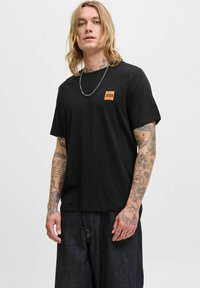 Schwarzes Baumwoll-T-Shirt mit kurzen Ärmeln, versehen mit einem kleinen orangefarbenen Quadrat-Logo auf der Brust. Modell trägt dunkle Jeansshorts.
