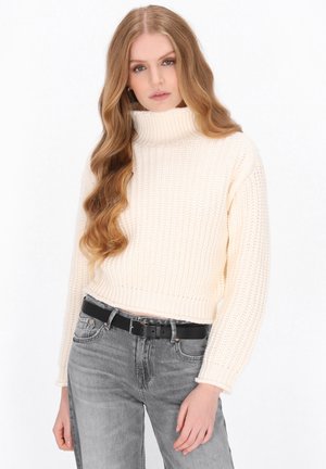 Donna con capelli lunghi e ondulati che indossa un maglione a collo alto color crema e jeans grigi sbiaditi con una cintura nera, in piedi davanti a uno sfondo bianco.