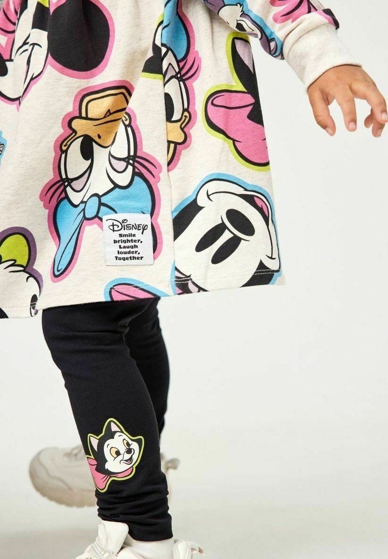 Enfant portant une robe et des leggings avec des imprimés colorés de personnages de dessins animés Disney, dont Donald Duck et un chat souriant, debout sur un fond blanc.