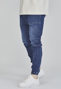 Blå denimjoggers med elastiska muddar och sidfickor, kombinerat med vita sneakers. Materialet verkar slätt med lätt bleknade detaljer.