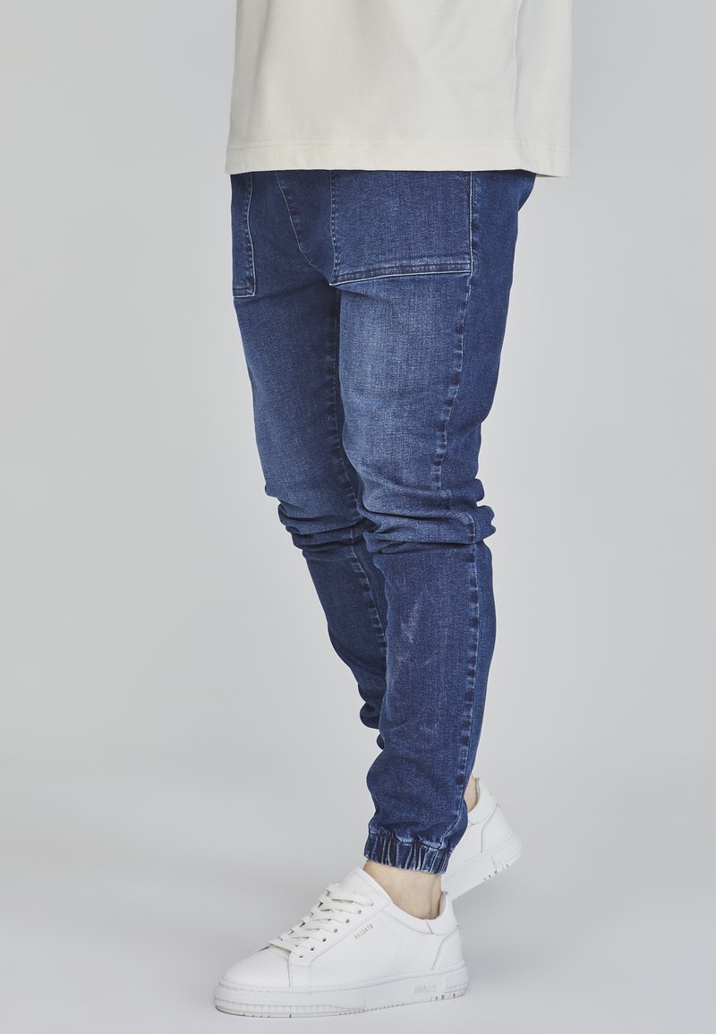 Blå denimjoggers med elastiska muddar och sidfickor, kombinerat med vita sneakers. Materialet verkar slätt med lätt bleknade detaljer.