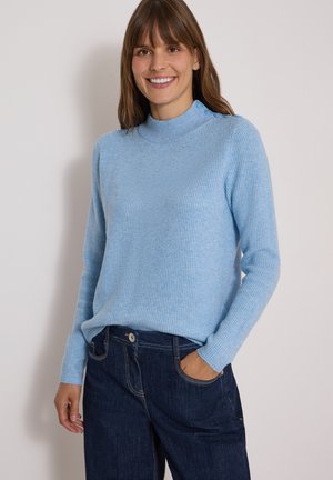 Smilende kvinde iført lyseblå strikket sweater med skulderknapper og mørkeblå jeans, stående mod en ensartet lys baggrund.