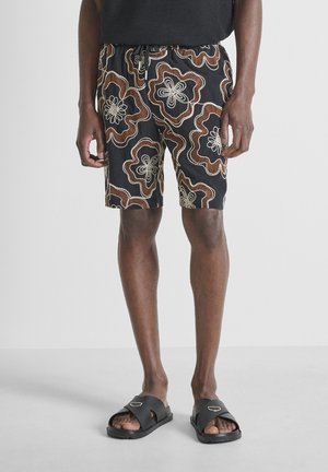 Uomo che indossa pantaloncini neri con coulisse e motivo floreale marrone e bianco, sandali neri tipo slide, in piedi su pavimento grigio chiaro.