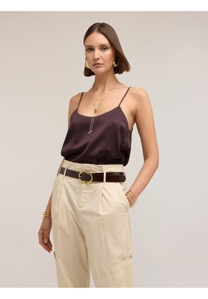 Femme portant un caraco en satin violet foncé, un pantalon beige taille haute avec une ceinture foncée, des boucles d'oreilles en or, des bracelets et un long collier.