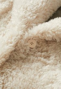 Beige texturovaná tkanina s nadýchaným vzhledem, obsahující světle béžový knoflík se čtyřmi dírkami. Detailní pohled na měkký, chlupatý materiál.