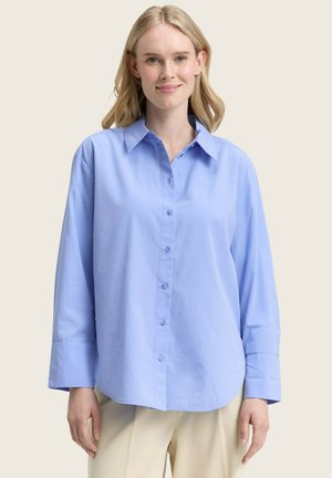Frau, die ein hellblaues Button-Up-Hemd mit langen Ärmeln und beigen Hosen trägt, steht vor einem schlichten hellen Hintergrund und lächelt leicht.