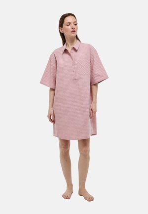 Femme debout pieds nus portant une robe chemise ample rose à carreaux avec manches courtes, une poche avant et des boutons, sur un fond uni.