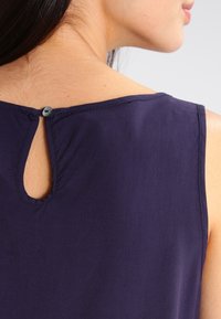 Navyblå ärmlös topp med en nyckelhålsöppning bak, fäst med en blå knapp. Slät textur och enkel design.