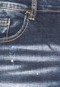 Jeans de mezclilla azul oscuro con patrones de desvanecimiento ligero, costuras doradas en contraste, diseño de cinco bolsillos y manchas de pintura blanca.