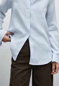 Camicia azzurro chiaro, in tessuto testurizzato, con colletto a punta, maniche lunghe e orlo curvo. Abbinata a pantaloni marroni.