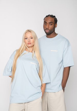 Two adults wearing light blue oversized shirts and beige pants stand side by side against a plain background.