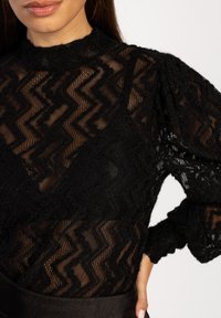 Schwarzes, durchscheinendes Oberteil mit Zickzackmuster, strukturiertem Strick, hohem Kragen, langen Puffärmeln und tailliertem Schnitt. Der Hintergrund ist weiß.