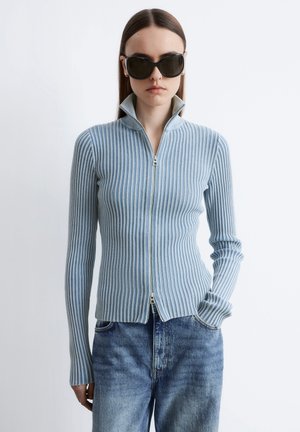 Kvinde iført sorte, store solbriller, en lyseblå stribet trøje med lynlås og blå højtaljede jeans, stående med den ene hånd i lommen.