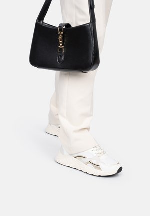 Baskets de sport blanches avec des mailles et des accents dorés, associées à un pantalon de couleur crème. Un sac à main noir avec un fermoir doré repose sur l'épaule.
