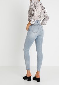 Kvinna i ljusblå skinny jeans, en långärmad blus med ormskinnsmönster och svarta klackskor med blockklack, som står mot en vit bakgrund.