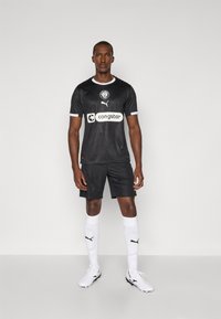 Maillot de football noir à manches courtes et col rond, avec des accents blancs. Comprend le logo du sponsor et la marque Puma. Porté avec des chaussettes blanches.