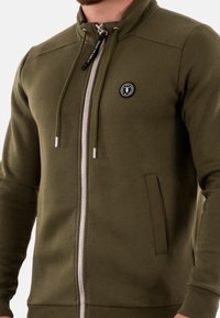 Giacca zip-up verde oliva con collo alto, un patch rotondo con logo, tasche laterali e zip a contrasto. Tessuto morbido con una superficie liscia.