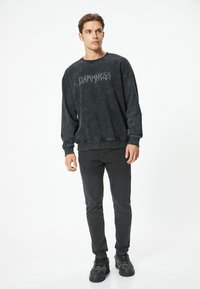 Sudadera negra de cuello redondo con un acabado texturizado, que presenta la palabra "OSCURIDAD" en una fuente estilizada. Combinada con unos vaqueros negros ajustados y zapatillas.