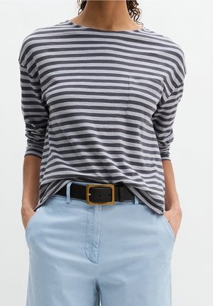 Personne portant une chemise à manches longues rayée grise et noire rentrée dans un pantalon bleu clair avec une ceinture noire et une boucle dorée, les mains dans les poches.
