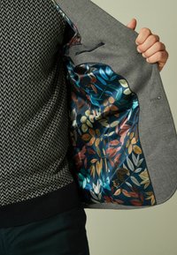 Grau jacket mit Hahnentrittmuster, ausgestattet mit einem bunten Blumenmuster-Futter in Blau-, Orange- und Grüntönen. Strukturierter Stoff.