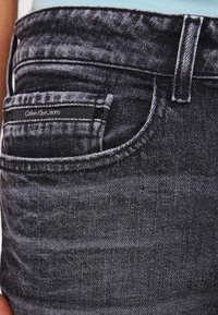 Vaqueros de mezclilla color índigo oscuro con un pequeño bolsillo delantero y una etiqueta de Calvin Klein Jeans. La tela tiene una textura sutil y un ligero desgaste.