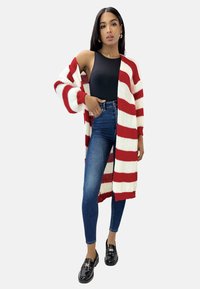 Czerwono-biały sweter w paski na drutach z otwartym przodem, zestawiony z czarnym bezrękawnym body i niebieskimi dżinsami z wysokim stanem. Czarne buty.