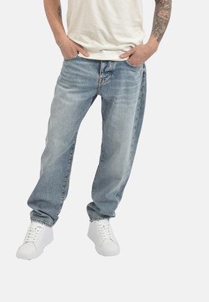 Man draagt lichtblauwe losse spijkerbroek, witte sneakers en een wit T-shirt, handen in de zakken, toont getatoeëerde onderarm.