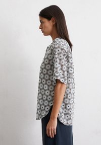 Blouse à manches courtes en tissu doux, présentant un motif floral répétitif en blanc, marine et marron clair, avec un ourlet arrondi.