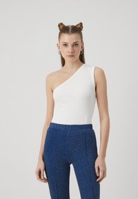 Hvit ribbet top med én skulder i kombinasjon med blå glitrende høytlivsbukser. Toppen har en figursydd design, mens buksene har sidesømmer.