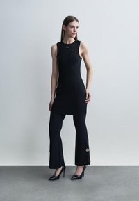 Γυναίκα στέκεται φορώντας μαύρο μίνι φόρεμα χωρίς μανίκια με μικρές διακοσμητικές λεπτομέρειες πάνω από μαύρο παντελόνι με καμπάνα και κουμπιά, μαύρα τακούνια, ουδέτερο φόντο.