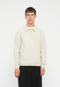 Suéter de punto beige con cuello, que presenta un dobladillo y puños acanalados, usado sobre una camisa blanca, combinado con pantalones oscuros de cuadros.