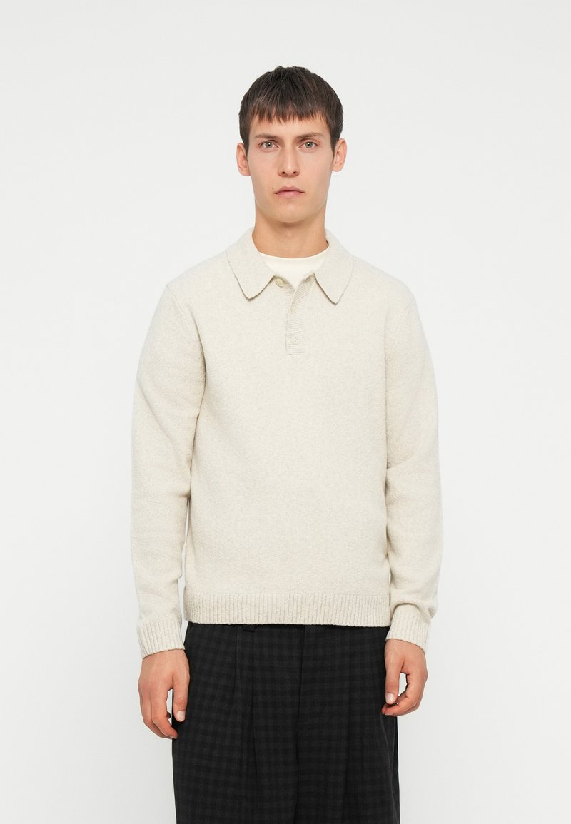 Suéter de punto beige con cuello, que presenta un dobladillo y puños acanalados, usado sobre una camisa blanca, combinado con pantalones oscuros de cuadros.