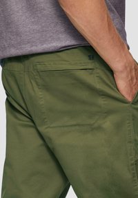 Pantalon vert olive en tissu résistant, avec deux poches arrière et un détail de logo au-dessus de la poche droite. Texture lisse et design structuré.