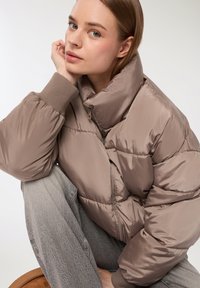 Giacca taupe morbida con colletto alto, polsini a coste e chiusura con zip; superficie testurizzata con cuciture visibili, indossata sopra pantaloni grigi.