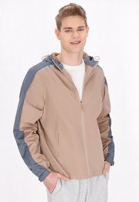 Jachetă ușoară de culoare bej cu accente gri, cu închidere prin fermoar, glugă ajustabilă și buzunare laterale. Textură netedă, design sportiv și croială modernă.