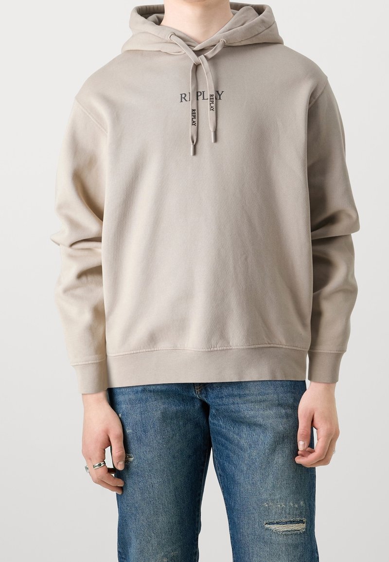 Personne porte un sweat à capuche beige avec le texte « REPLAY » et des cordons, ainsi qu’un jean bleu délavé, debout devant un fond clair et neutre.