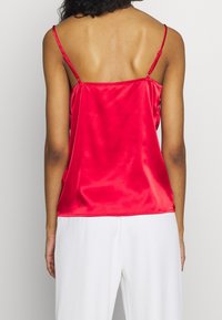 Rød satin camisole med tynde stropper, glat tekstur og en afslappet pasform, der hænger blødt over ryggen, parret med hvide bukser.