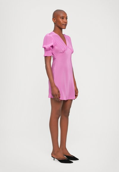 Korte roze satijnen jurk met pofmouwen en een diepe V-hals. Model heeft het gecombineerd met zwarte instapheels. Gladde textuur en knielange ontwerp.