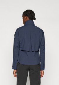 Mörkblå, långärmad jacka med hög krage. Har en plisserad bakdesign och justerbara ärmslut, tillverkad av ett lätt material.