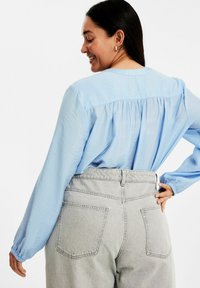 Hellblaue Bluse mit langen Ärmeln und gerafftem Rücken, kombiniert mit hoch taillierten grauen Jeans mit Gesäßtaschen und einer lockeren Passform.