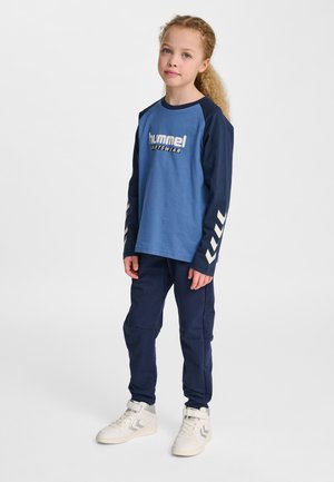 Blå langærmet skjorte med marineblå ærmer og hvidt logo, kombineret med marineblå bukser og hvide sneakers med grå accenter.